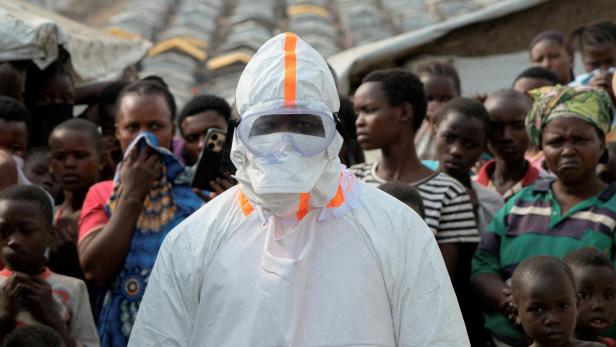 Eine Person in weißer Schutzkleidung mit Maske und Schutzbrille steht vor einer Menschenmenge in einem Lager mit zahlreichen Zelten.