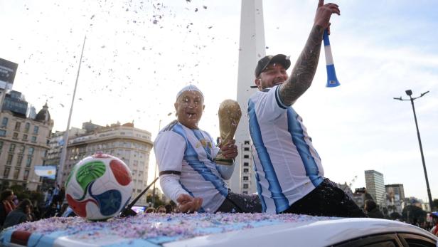 Zwei Fans in Argentinien-Trikots feiern mit dem WM-Pokal vor dem Obelisken in Buenos Aires inmitten von Konfetti.