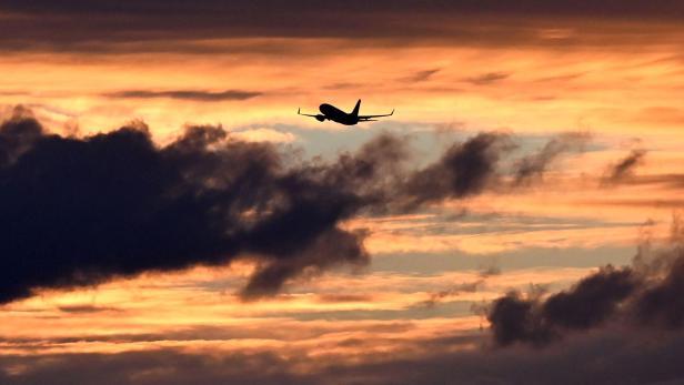 Die Silhouette eines Flugzeugs fliegt vor einem dramatischen, orangefarbenen Sonnenuntergangshimmel mit dunklen Wolken.