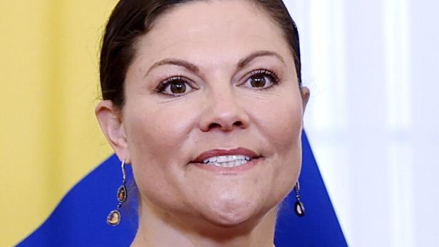 Kronprinzessin Victoria von Schweden trägt ein dunkelrotes Oberteil vor der schwedischen Flagge.