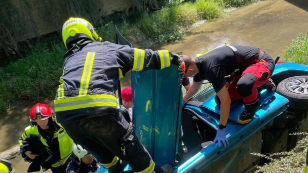 Feuerwehrkräfte retten eine Person aus einem auf der Seite liegenden blauen Auto am Bachufer.