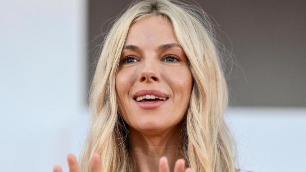 Sienna Miller lächelt mit erhobenen Händen und trägt ein weißes Spitzenoberteil mit einem auffälligen goldenen Chloé-Gürtel.