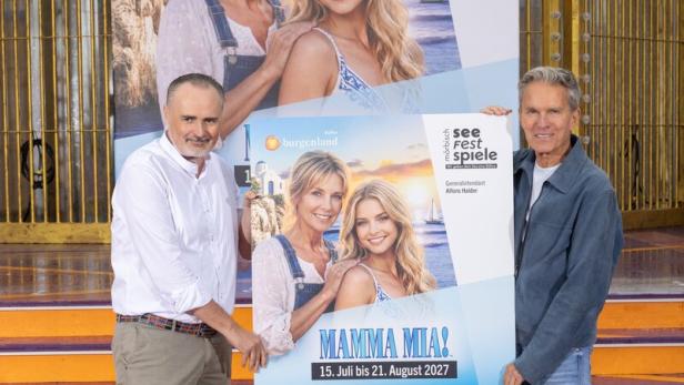 Zwei Männer halten ein Mamma-Mia!-Plakat vor einer goldenen Kulisse.