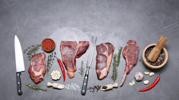 Vier rohe Steaks mit Messern, Kräutern, Knoblauch, Chilischoten und Gewürzen auf grauem Hintergrund.
