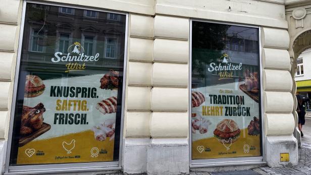 Schaufenster eines Schnitzel-Lokals mit Werbeplakaten für Burger, Hähnchen und Slogans wie „Knusprig. Saftig. Frisch.“.