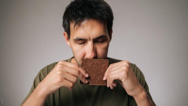 Studio portrait of enjoying young man savoring rich aroma of dark chocolate bar with closed eyes, holding it close to face in anticipation of delightful treat
