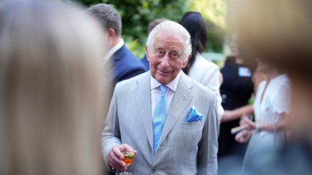 König Charles III. lächelt in einem hellen Anzug mit blauem Einstecktuch und hält ein Glas in der Hand, umgeben von Gästen bei einer Veranstaltung.