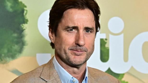 Luke Wilson trägt ein hellblaues Hemd unter einem beigen Sakko vor einem unscharfen Hintergrund.