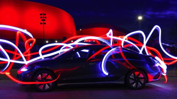 Ein schwarzer Elektro-Pkw steht vor einem rot beleuchteten Gebäude, umgeben von roten und weißen Lichtspuren.