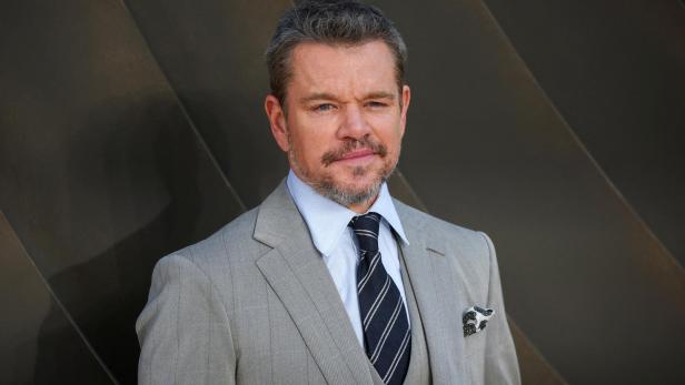 Matt Damon trägt einen grauen Anzug mit Krawatte vor einem dunklen, geometrisch strukturierten Hintergrund.