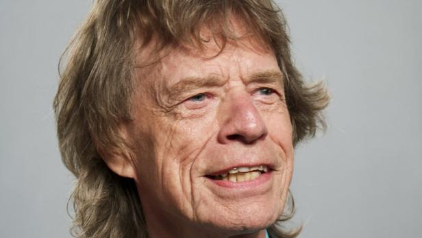 Mick Jagger lächelt vor einem grauen Hintergrund in einem türkisfarbenen Hemd und einem gemusterten Sakko.