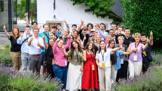 Eine große Gruppe junger Menschen steht vor einem Gebäude im Freien und zeigt gemeinsam den Daumen nach oben.