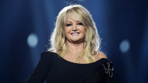 Bonnie Tyler lächelt vor einem dunklen, blau beleuchteten Hintergrund in die Kamera.