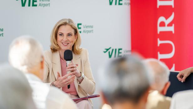 Eine blonde Frau spricht vor Publikum in ein Mikrofon vor einer Wand mit dem Logo des Vienna Airport.
