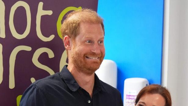 Prinz Harry lächelt neben einer Krankenschwester der Wohltätigkeitsorganisation WellChild vor einem Werbebanner.