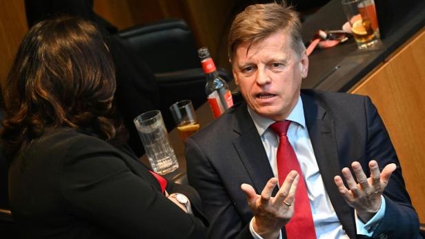 Ein Mann im Anzug mit roter Krawatte gestikuliert lebhaft während eines Gesprächs mit einer Frau in einem Konferenzraum.