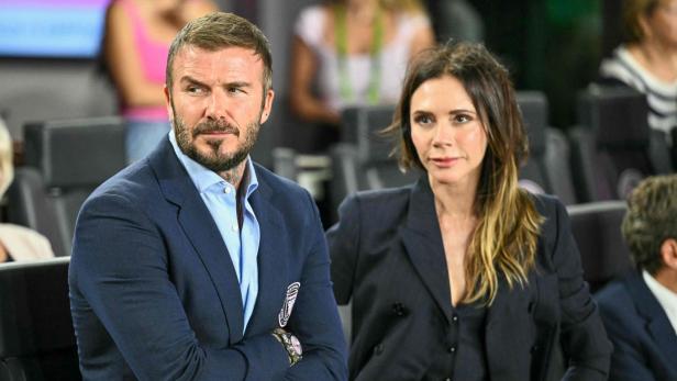 David und Victoria Beckham sitzen nebeneinander in eleganten dunklen Anzügen auf einer Tribüne.