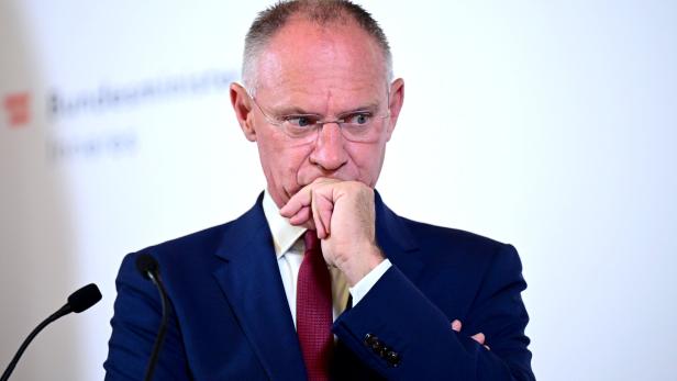 Ein Mann im Anzug und mit Brille steht nachdenklich an einem Rednerpult, die Hand vor dem Mund.