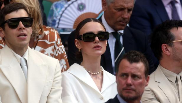 Keira Knightley trägt eine Sonnenbrille und ein weißes Oberteil, während sie neben anderen Zuschauern auf der Tribüne in Wimbledon sitzt.