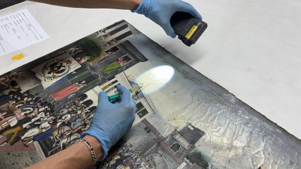 Mit Handschuhen wird ein altes Gemälde mit Lampe und Werkzeug untersucht.