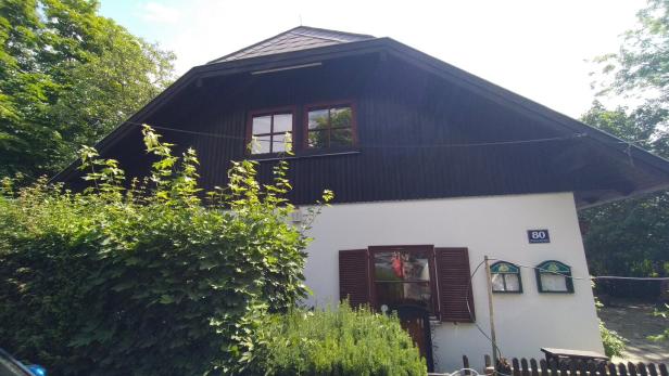 Zweistöckiges Haus mit dunklem Dach, weißen Wänden und viel Grün davor, im Vordergrund ein Zaun.