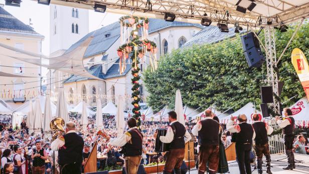 Eine Musikkapelle in Tracht spielt auf einer Bühne vor einem Maibaum und einer Menschenmenge auf einem Marktplatz.