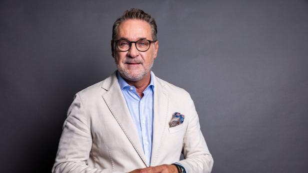 Heinz Christian Strache mit Brille, grauem Bart und hellem Sakko steht vor einem grauen Hintergrund.
