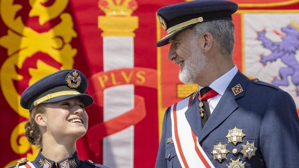 König Felipe VI. von Spanien lächelt seiner Tochter Prinzessin Leonor in Militäruniform vor einer spanischen Flagge zu.