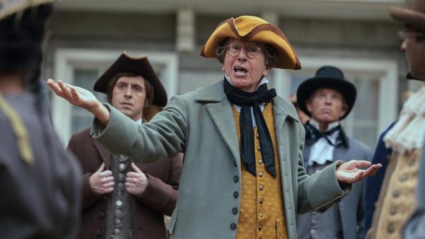 Ein Mann (Larry David) mit Brille und in historischer Kleidung mit Dreispitz gestikuliert lebhaft vor einer Gruppe weiterer Personen in zeitgenössischen Kostümen.
