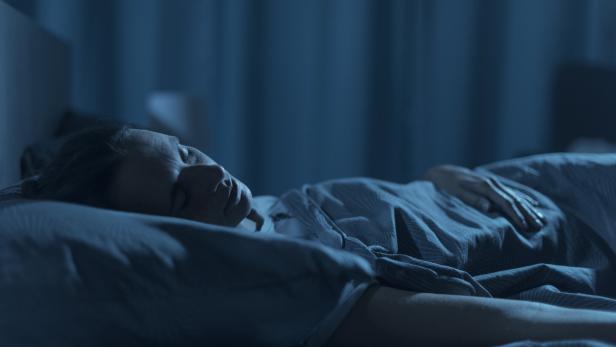 Eine schlafende Person liegt im Bett, nachts und blau beleuchtet, die Hand auf dem Bauch.