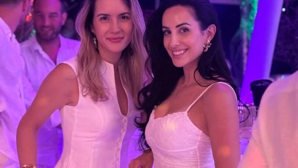 Zwei Frauen in weißen Outfits lächeln bei einer Party im violetten Licht.