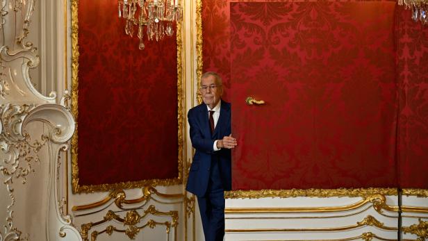 Der österreichische Bundespräsident Alexander Van der Bellen steht in einem prunkvollen Raum mit roter Tapete hinter einer halb geöffneten Tür.