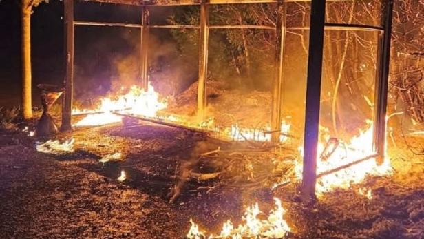 Ein offener Unterstand aus Metall steht in Flammen, mit Feuer auf dem Boden und an den Pfosten.
