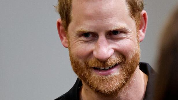 Prinz Harry lächelt freundlich in einem schwarzen Poloshirt vor einem neutralen Hintergrund.