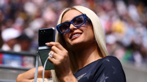 Paris Hilton macht mit einem Smartphone ein Selfie vor einer unscharfen Zuschauerkulisse.