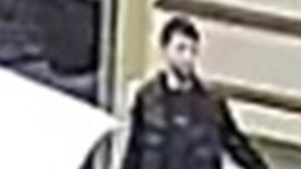 Mann in dunkler Jacke und Jeans steht neben einem Auto an einer Hauswand.