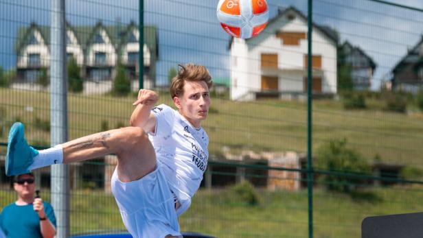 Ein junger Mann in weißer Sportkleidung spielt Teqball auf einem blauen Außenplatz.