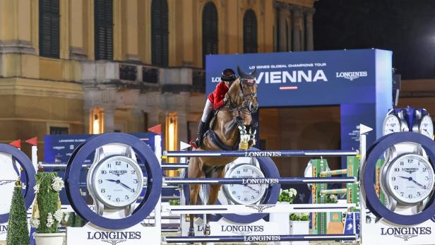 Springreiter überwindet ein Longines-Hindernis bei Nacht vor historischer Fassade.