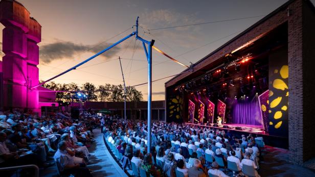 Open-Air-Theater bei Sonnenuntergang mit großem Publikum vor einer beleuchteten Bühne.