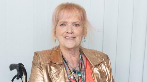 Gaby Köster mit blondem Haar trägt eine goldene Jacke und viele Halsketten, während sie in einem Rollstuhl sitzt.