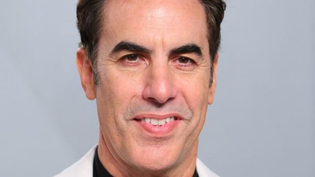 Sacha Baron Cohen lächelt vor einem grauen Hintergrund in einem weißen Sakko über einem schwarzen Rollkragenpullover.