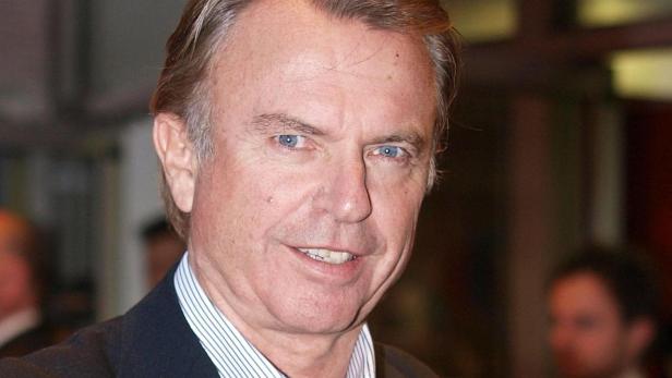 Der Schauspieler Sam Neill lächelt in einem dunklen Sakko und einem gestreiften Hemd vor verschwommenem Hintergrund.