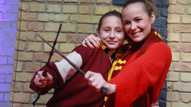 Zwei grinsende Personen in Gryffindor-Kleidung umarmen sich und halten Zauberstäbe vor einer Backsteinwand.