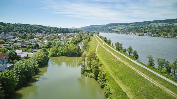 Luftaufnahme eines Flusstals mit Damm, Uferweg, Wohnhäusern und bewaldeten Hügeln.