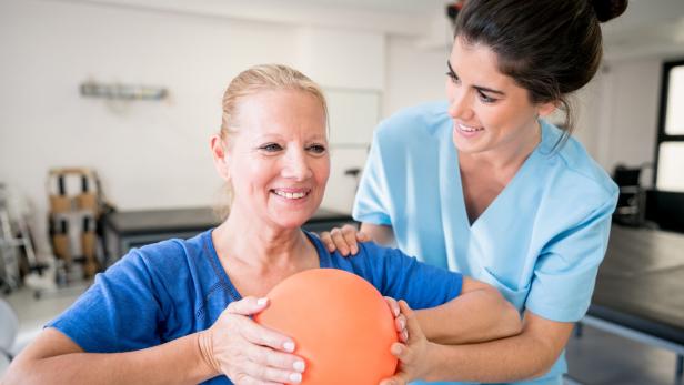 Physiotherapeutin unterstützt lächelnde Patientin beim Halten eines orangefarbenen Balls.