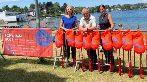 Drei Personen stehen am See vor einer Reihe orangefarbener Schwimmbojen und einem KFV-Banner.