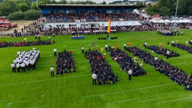 Zahlreiche Feuerwehrleute in Uniform sitzen in Gruppen auf einem Sportplatz vor einer voll besetzten Tribüne bei einer feierlichen Veranstaltung.
