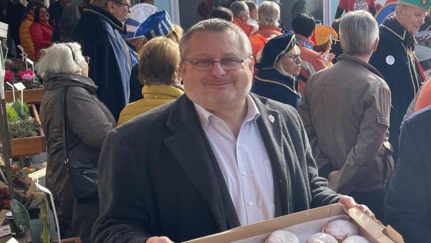 Mann lächelt in die Kamera und hält eine Kiste mit Puderzucker-Krapfen auf einem vollen Festplatz.