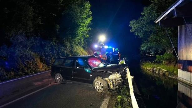 Nächtliche Unfallstelle: Ein schwarzer Kombi mit Frontschaden steht neben der Leitplanke, Rettungskräfte sind vor Ort.