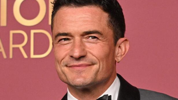 Orlando Bloom lächelt vor einem roten Hintergrund in einem grauen Smoking mit schwarzem Revers und einer Fliege.